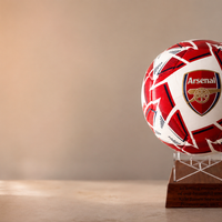 An Arsenal soccer ball with a red and white pattern, displayed on a clear acrylic stand atop a wooden plaque, set against a warm, softly lit indoor background.