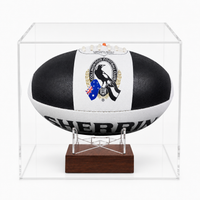 A black and white AFL ball with a Magpies logo, displayed inside a clear acrylic case on a wooden stand.