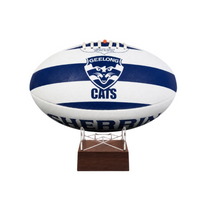 A white and navy blue Geelong Cats  AFL ball displayed on a small wooden stand against a plain white background.