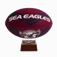 A maroon and navy blue rugby ball with 'SEA EAGLES' in white text and the Manly Warringin club logo, displayed on a clear acrylic stand with a dark wood base against a white background.