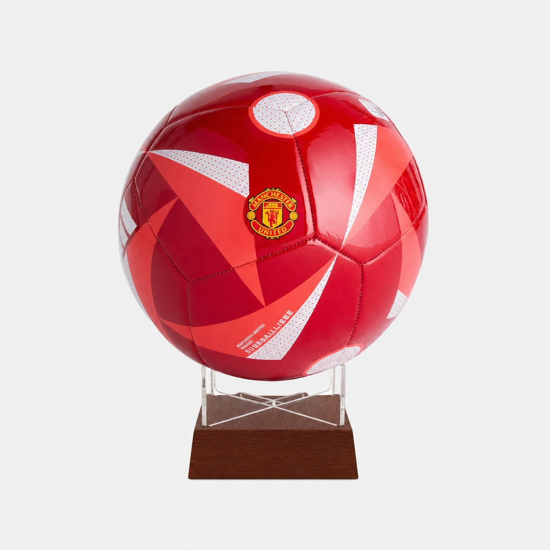 A red and white Manchester United soccer ball mounted on a clear acrylic stand with a dark brown wooden base, set against a light gray background.